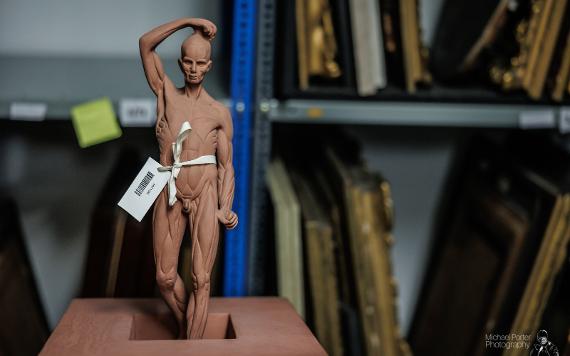 Small statue of man tagged up inside Harris storage room, with paintings on shelves in background.