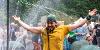 A performer stands under a shower of water with ripped up clothes and his arms outstretched in joy