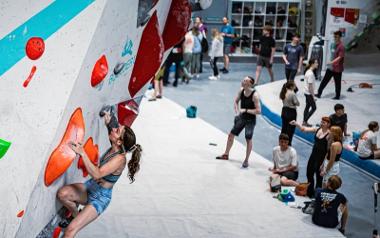 A crowd of people wait to climb an indoor climbing wall as they watch a lady climbing