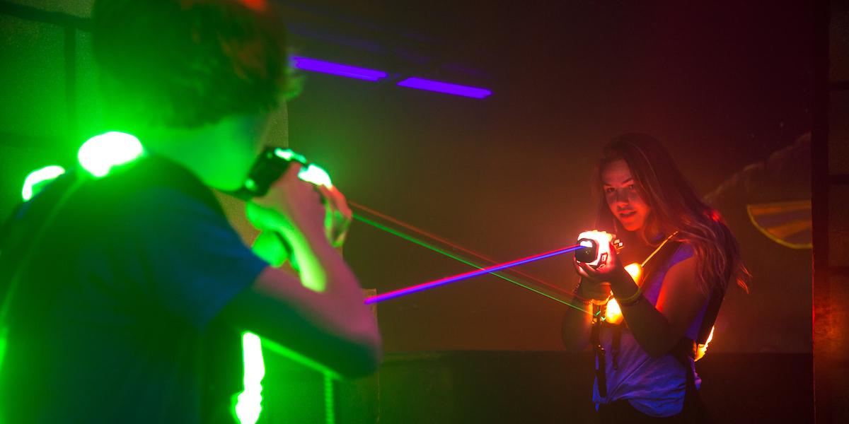 A girl with an orange laser gun faces a boy with a green laser gun inside Laser Quest arena.