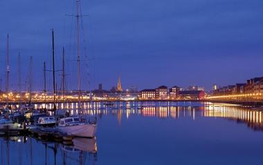 Boats, skyline, and lights reflected in the water of Preston Dock at night.