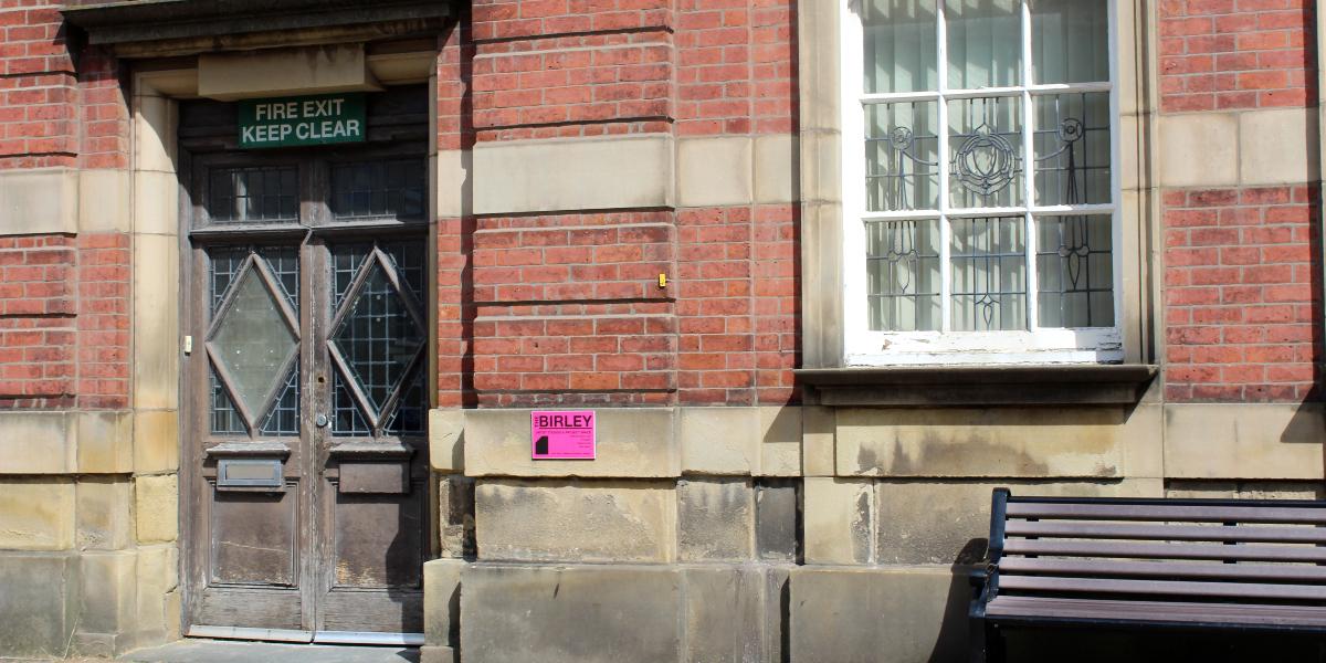 Front entrance of The Birley building with bench beside door.