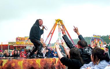 Singer on Rockprest stage with crowds and fairground rides in background.